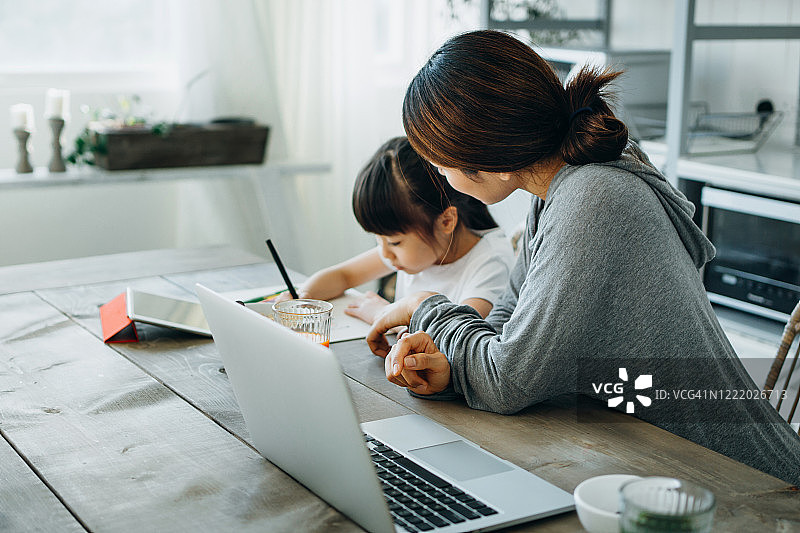年轻的亚洲母亲在家用笔记本电脑工作。在家教育她的女儿，并在家里帮助她完成学业图片素材