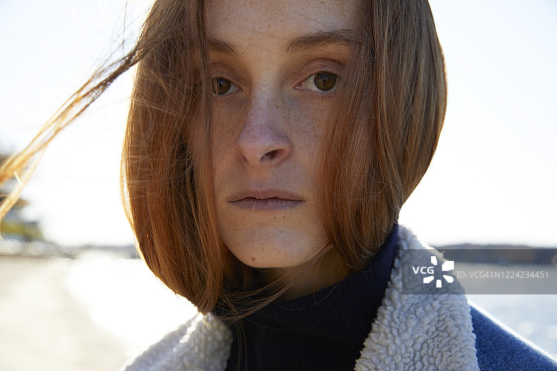 一个漂亮的红发少女的肖像，在外面的海滩上。看着摄像机。她有一双棕色的眼睛，风吹着她的头发。图片素材