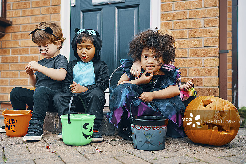 三个穿着万圣节服装的孩子坐在门阶上吃糖果。图片素材