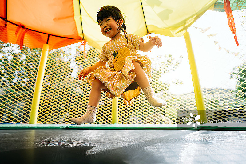 快乐的小亚洲学步的女孩愉快地微笑，并有乐趣在一个充气城堡在户外操场上跳跃图片素材