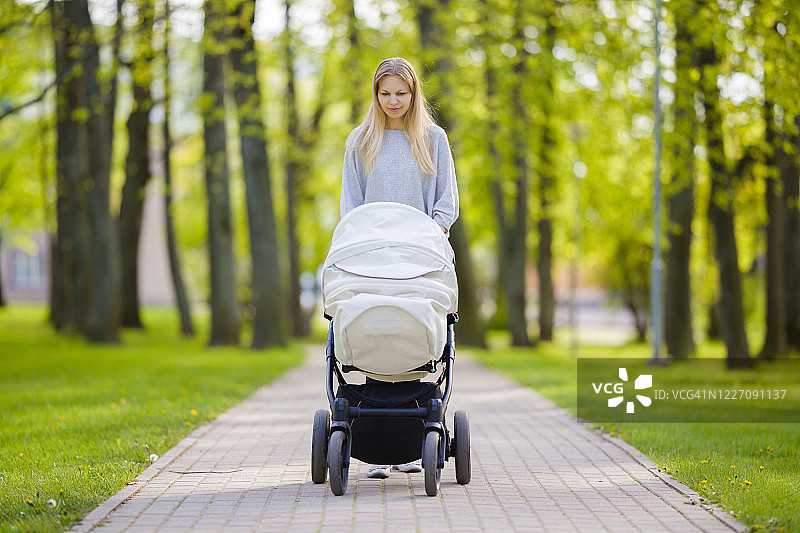一位年轻的母亲推着白色婴儿车，在温暖、阳光明媚的夏日小镇绿地公园慢慢地走着。花时间陪婴儿，呼吸新鲜空气。享受漫步。前视图。图片素材