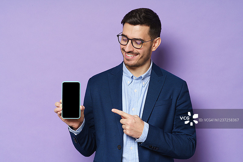 年轻的商人穿着夹克、衬衫和眼镜，拿着空白屏幕的智能手机，指着它，建议高质量的金融应用，孤立在紫色的背景图片素材
