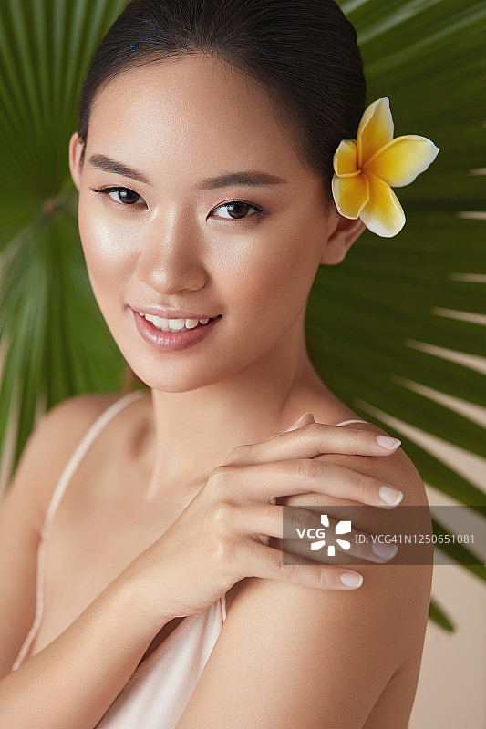 美。模型在棕榈与花的头发。温柔的亚洲女人触摸肩膀肖像在热带植物的背景。美丽的女孩享受水润肌肤后使用天然有机化妆品。图片素材