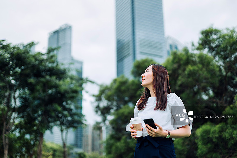 在市中心金融区的一个城市公园里休息时，体贴的年轻亚洲女商人端着智能手机，喝着咖啡，仰望着天空图片素材