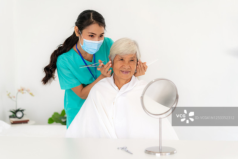 亚洲年轻女性护士或家庭健康助理梳头和剪头发与老夫妇在客厅餐桌在家的家庭保健概念。图片素材