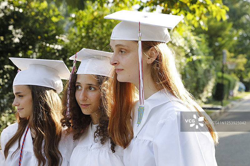 毕业日4名少女户外合影。他们都戴着白色的帽子，穿着白色的长袍，站在附近的街道上。美丽的背景光。图片素材