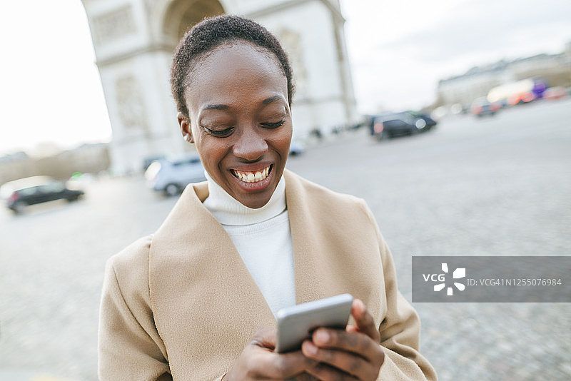 在阳光明媚的日子里，法国巴黎，一名快乐的年轻女子使用智能手机对抗凯旋门图片素材