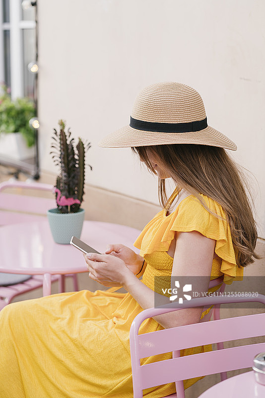 女人戴着帽子坐在城市路边咖啡馆的椅子上使用智能手机的侧视图图片素材