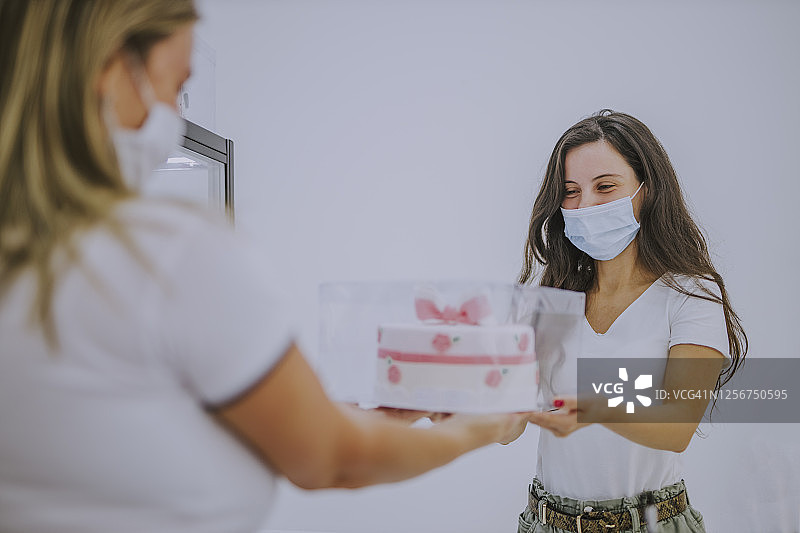 疫情期间，一名年轻女子戴着防护口罩，在她的蛋糕店递给顾客一个盒子图片素材
