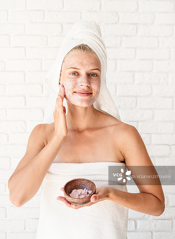 快乐微笑的年轻女子在白色浴巾应用磨砂在她的脸和颈部图片素材