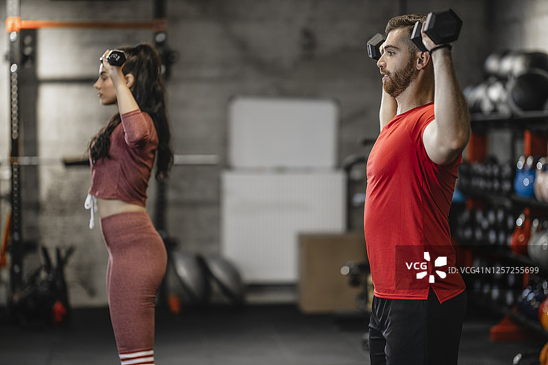 在年轻女性健身教练的帮助下，年轻的运动男子正在健身房用哑铃进行重量训练图片素材