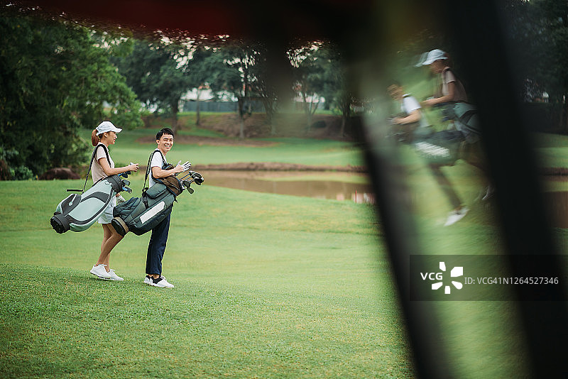 侧面的亚洲华人微笑年轻夫妇高尔夫球手拿着高尔夫球袋和站在高尔夫球场图片素材
