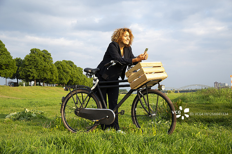 女商人一边用智能手机一边骑着自行车站在草地上，背靠天空图片素材