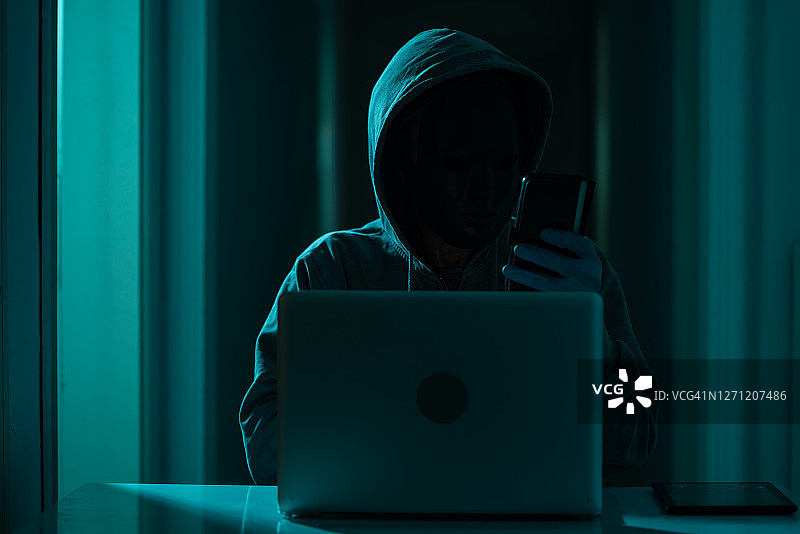 穿着连帽衫和面具的男子在黑暗中使用笔记本电脑进行黑客攻击图片素材