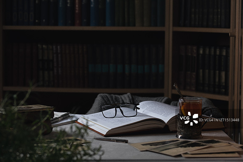 眼镜躺在打开的书和一杯茶。背景是书架图片素材