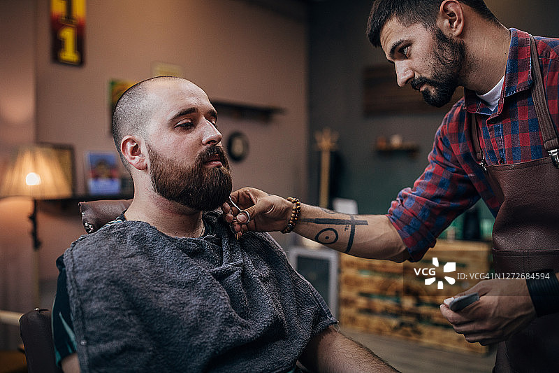 年轻的男性理发师在理发店用剪刀为顾客设计胡须图片素材