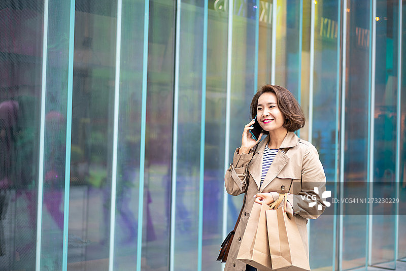 美丽的亚洲女性拿着购物袋，在城市街道上使用智能手机图片素材