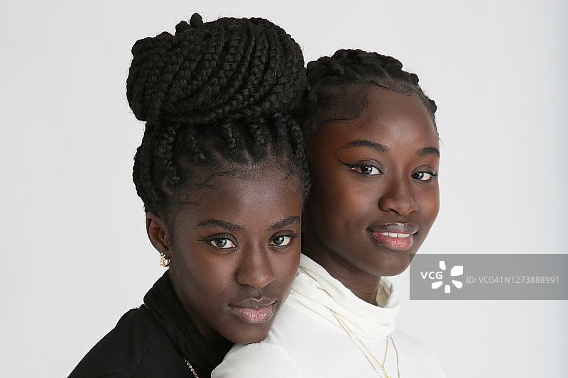 一对非洲血统双胞胎姐妹的肖像，穿着黑白高领上衣和牛仔裤，梳着长长的辫子，站在一个白色的工作室里。图片素材