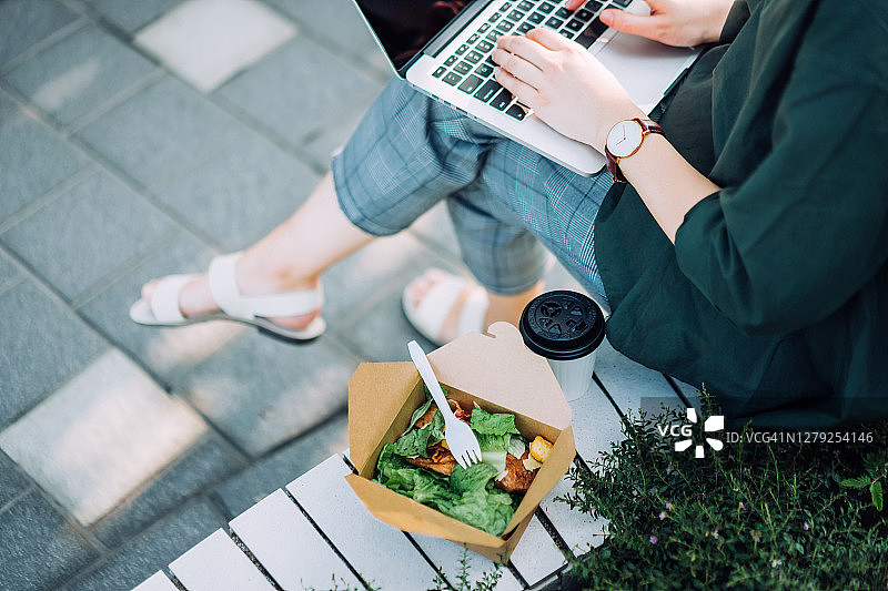 从头顶上看，一位年轻的亚洲女商人坐在城市公园的长椅上，一边用笔记本电脑工作，一边在午餐休息时间吃着健康的沙拉午餐盒和一杯咖啡图片素材