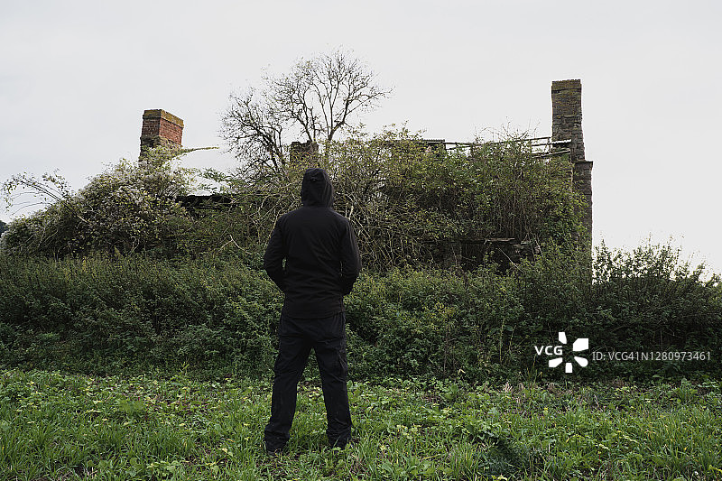 一座阴森森的废弃房子，上面覆盖着植被。一个戴兜帽的人站在乡下图片素材