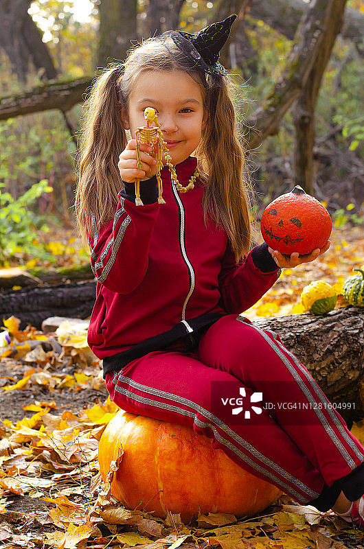欧洲女孩，头戴女巫帽，手里拿着骷髅和橙色南瓜，坐在一个大南瓜上。秋天的森林里有黄色的落叶。万圣节。图片素材