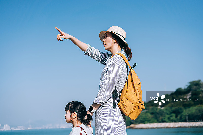 年轻的亚洲母亲牵着小女儿在大自然中徒步旅行。在蓝天和大海的映衬下，仰望天空。在一个阳光明媚的日子里，他们一起享受自然和美丽的风景图片素材