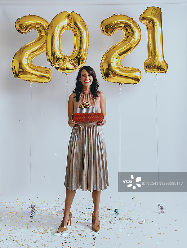 新的2021快乐!一个快乐的女人站在新年的气球下拿着礼物(看着相机)图片素材