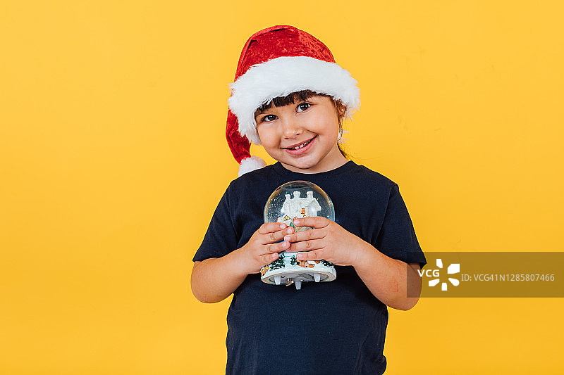 小女孩微笑着戴着圣诞帽，手里拿着圣诞雪花球，黄色的背景上。圣诞节的概念图片素材