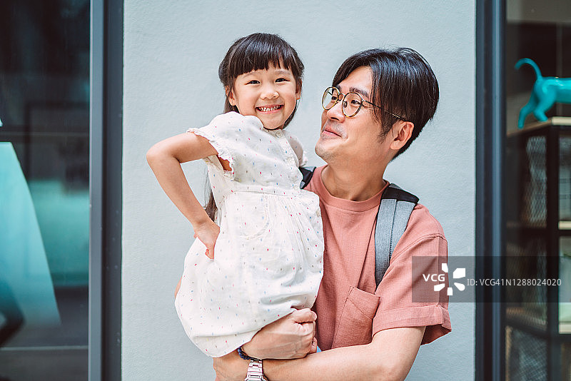 当她帅气的爸爸抱着她在购物中心时，可爱的小女孩对着镜头开心地笑图片素材