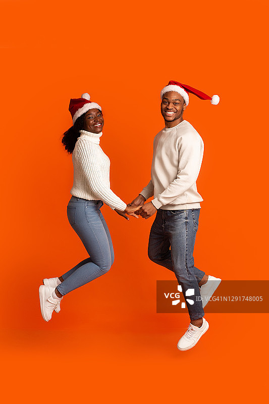 浪漫的黑人夫妇在圣诞老人帽子牵手和跳跃，橙色背景图片素材