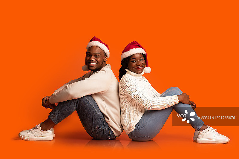 戴着圣诞老人帽的快乐黑人夫妇背靠背坐着的肖像图片素材