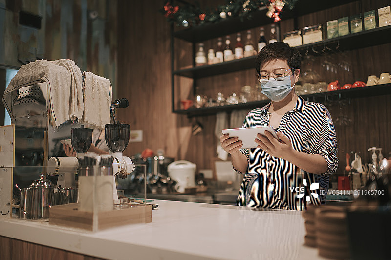 亚洲华人小企业主戴着口罩在厨房柜台微笑着看数字平板电脑图片素材
