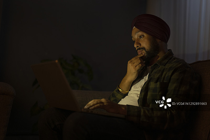 一名锡克教男子晚上一边用笔记本电脑工作一边思考图片素材