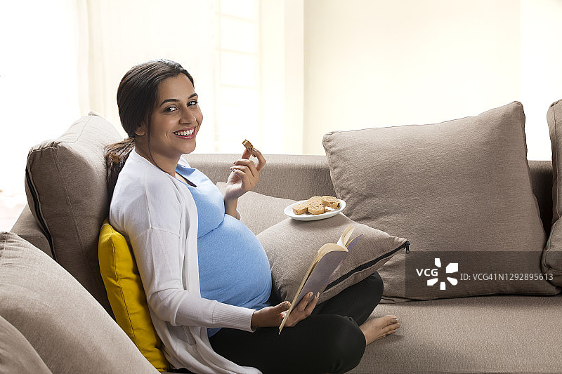 孕妇坐在沙发上一边吃一边看书。图片素材
