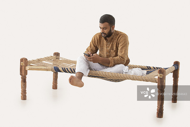 一个农村男子坐在椰壳床上使用手机的肖像图片素材