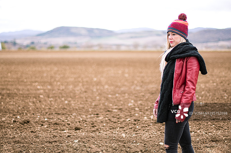一个红唇的金发女人正看着镜头，她穿着一件红色的皮夹克，一条黑色的围巾和一顶五颜六色的羊毛帽子。秋天，她走在新耕过的田地边上。图片素材