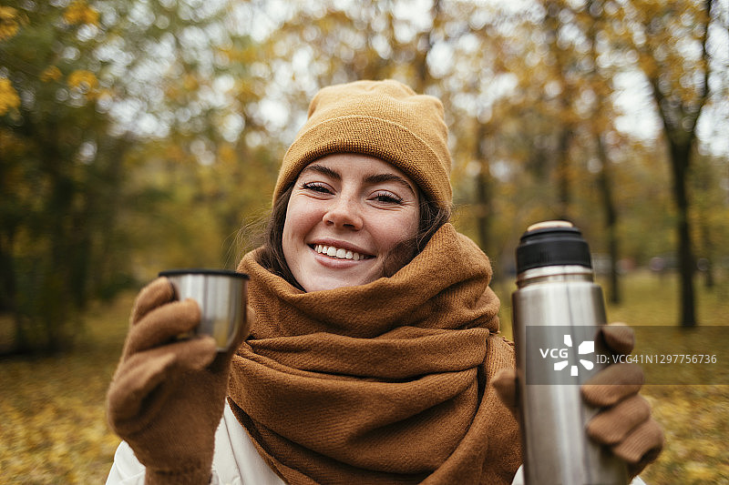 微笑的年轻女子与绝缘饮料容器在秋季公园图片素材