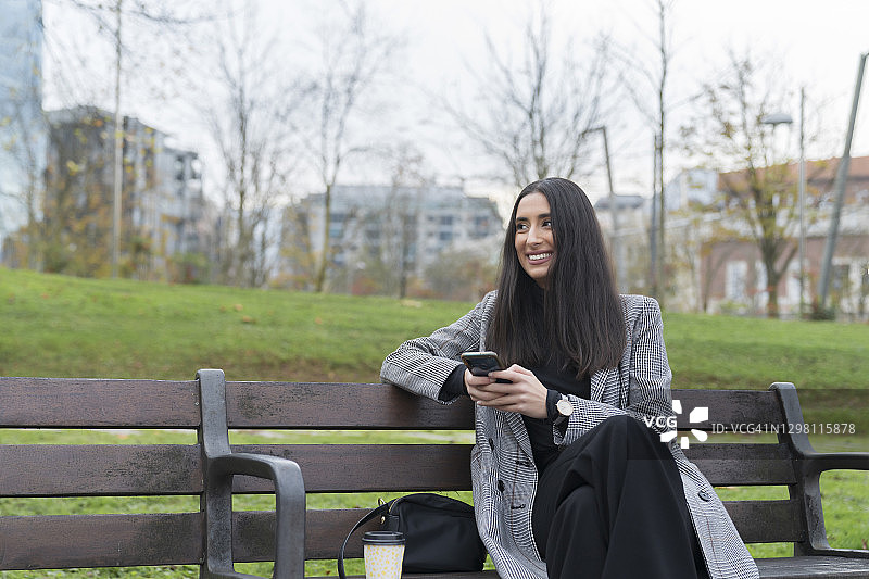 微笑的职业女性，拿着咖啡杯看向别处，在公园长凳上拿着手机图片素材