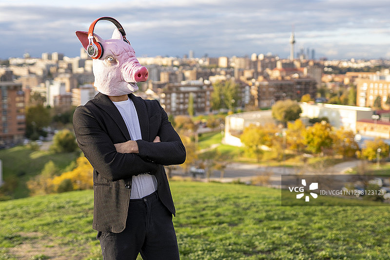 男企业家戴着猪面具和无线耳机，双臂交叉对着天空站着图片素材