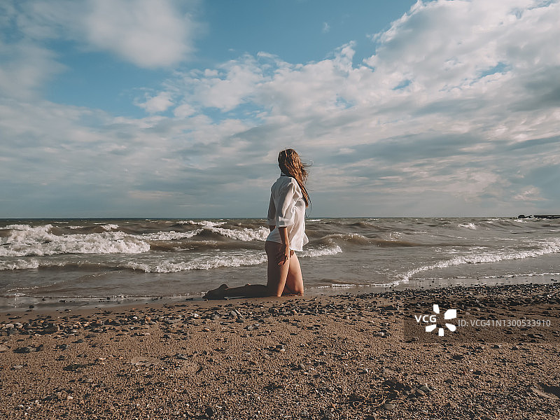 一个美丽的年轻女子与风吹头发在白色丝绸衬衫和比基尼在潮湿的沙滩上的海浪和暴风雨的天空的背景图片素材