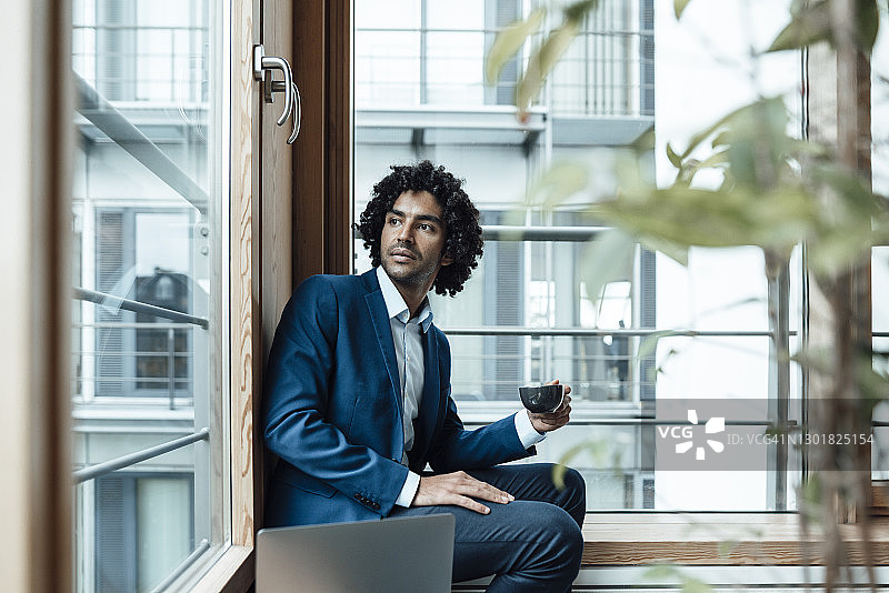在办公室里，体贴的年轻职业男性拿着咖啡杯，靠着窗户坐着笔记本电脑，把目光移开图片素材