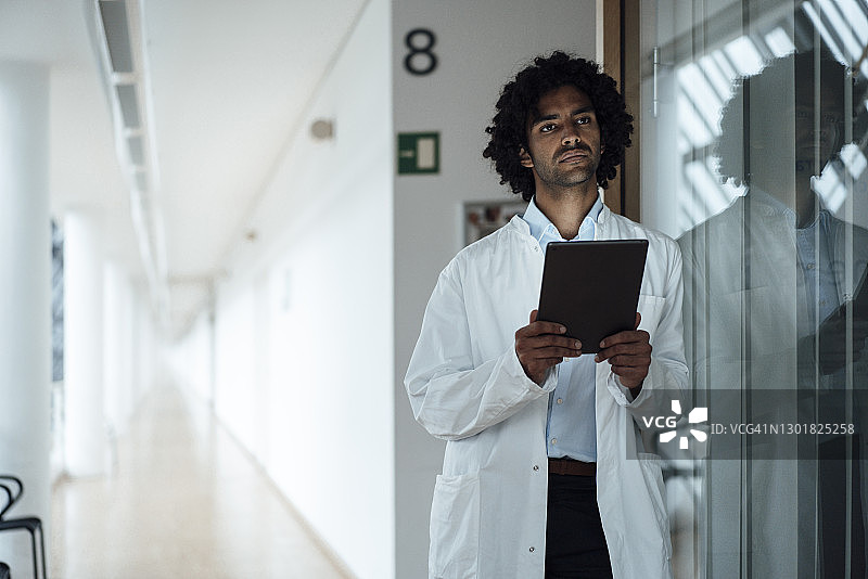 体贴的年轻男医生拿着电子平板电脑，倚靠在医院走廊的玻璃墙上图片素材