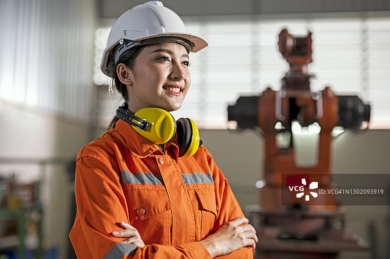 年轻女工业工程师的肖像与白色头盔抬头，而站在工业机器人前。工厂自动化和精益制造的概念。图片素材