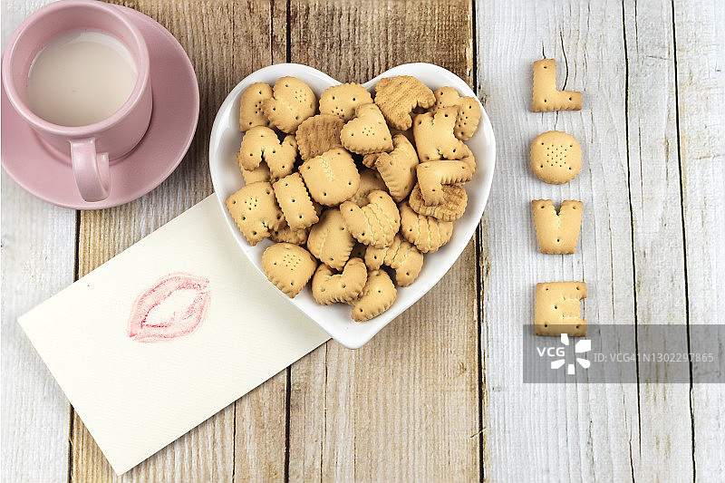 饼干的顶视图陶瓷心形碗，杯牛奶，字爱和信封上的口红亲吻与木材背景。图片素材