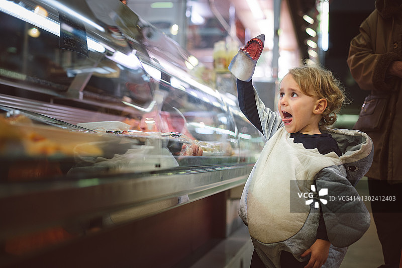 男孩打扮成鲨鱼在市场的肉区玩一个鲨鱼木偶，并伸出舌头图片素材