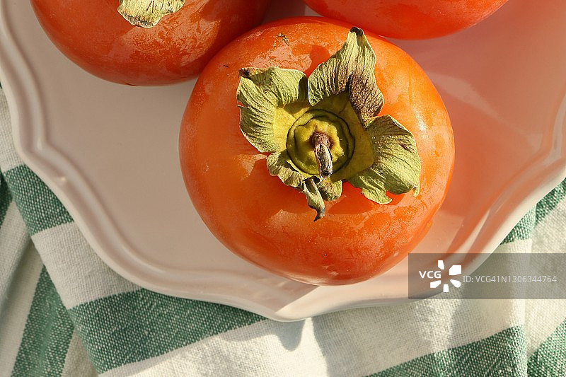新鲜成熟的柿子放在白色的盘子上，绿白相间的条纹桌布上，这是天堂棕榈图片素材