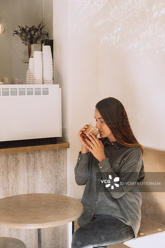 穿着绿色亚麻衬衫的年轻女子正在咖啡店的木桌后面喝咖啡。现代简约室内图片素材