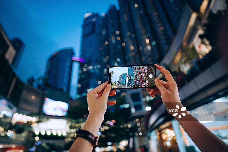 女性的手捕捉现代城市景观与高层金融建筑与照明的城市景观在晚上的智能手机的镜头图片素材
