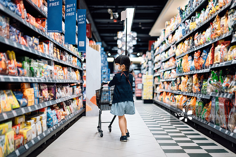 可爱的亚洲小女孩戴着保护面罩推着购物车，走在超市的过道上，购物图片素材