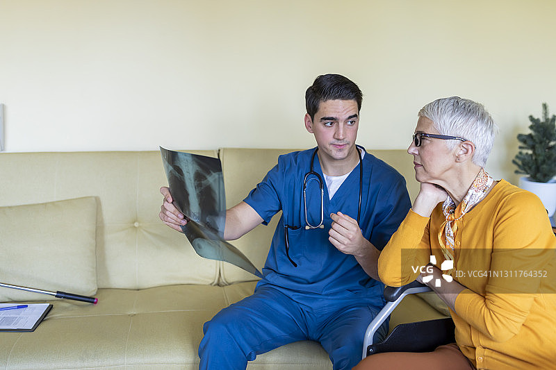 身穿蓝色制服的年轻男医生正在拜访一位坐在轮椅上的年长妇女，并在x光图像上诊断。图片素材